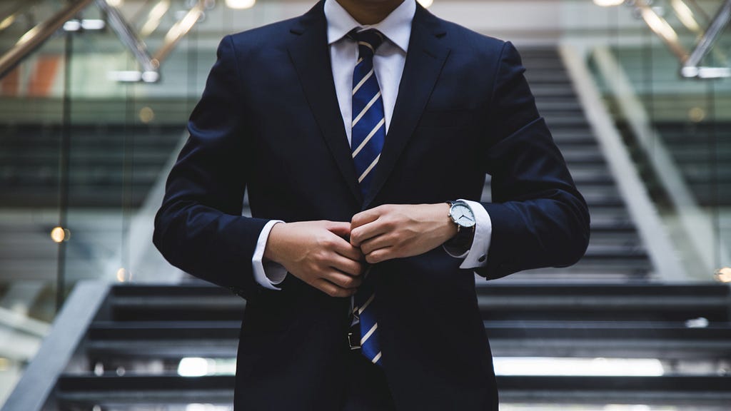 A white man in a navy suit adjusts his tie; his face is cropped