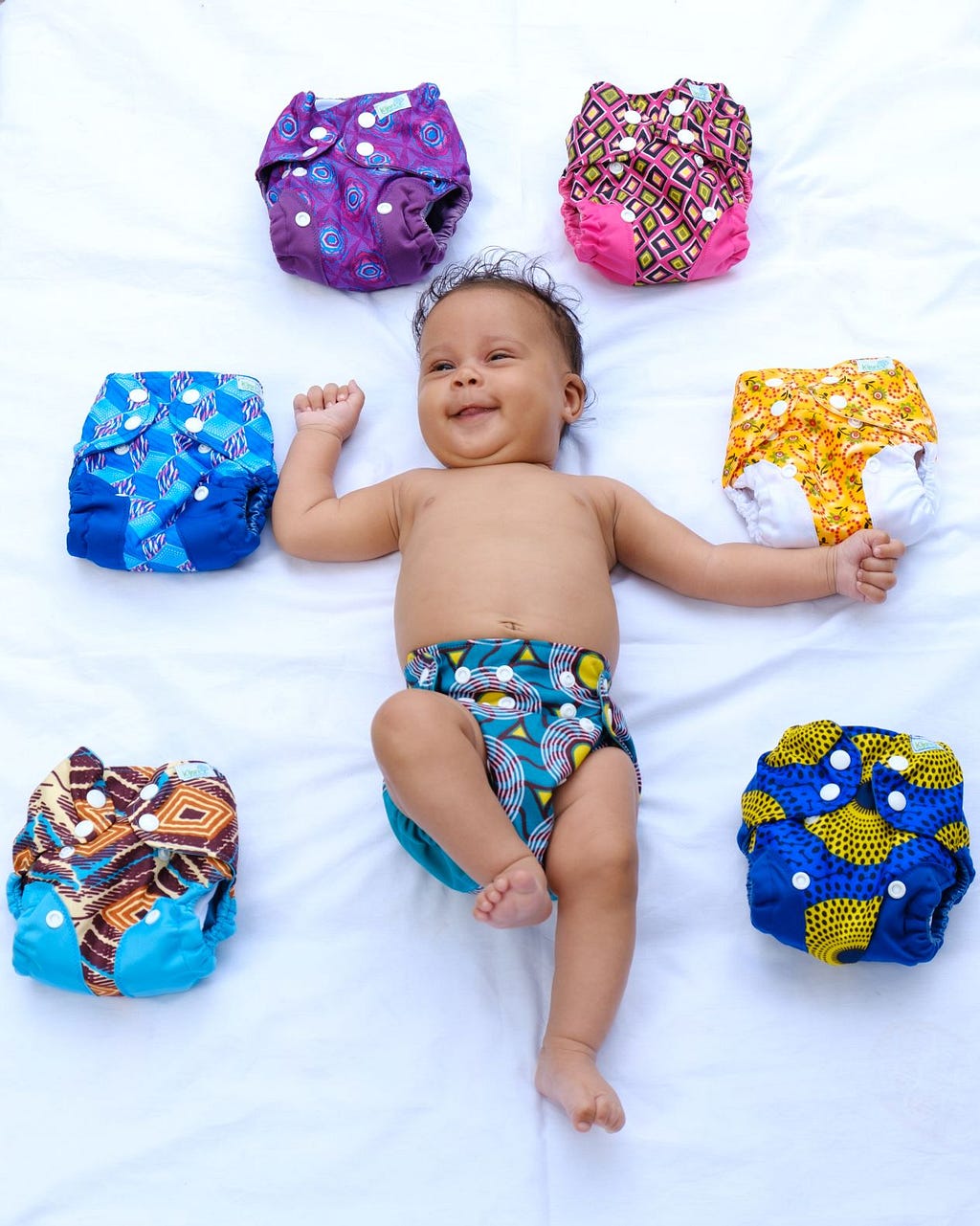 A photograph of a smiling baby, which lies on a white sheet surrounded by colourful nappies.