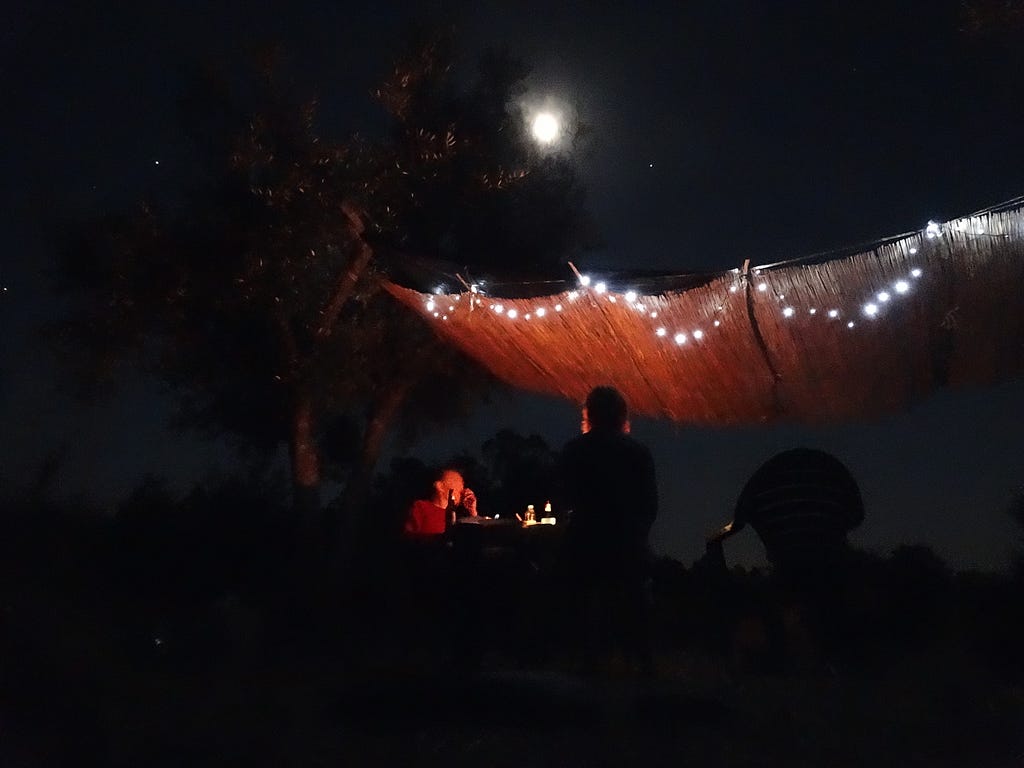Two people sit around a table on a moon-lit night beside an olive tree under a bamboo screen with fairy lights.