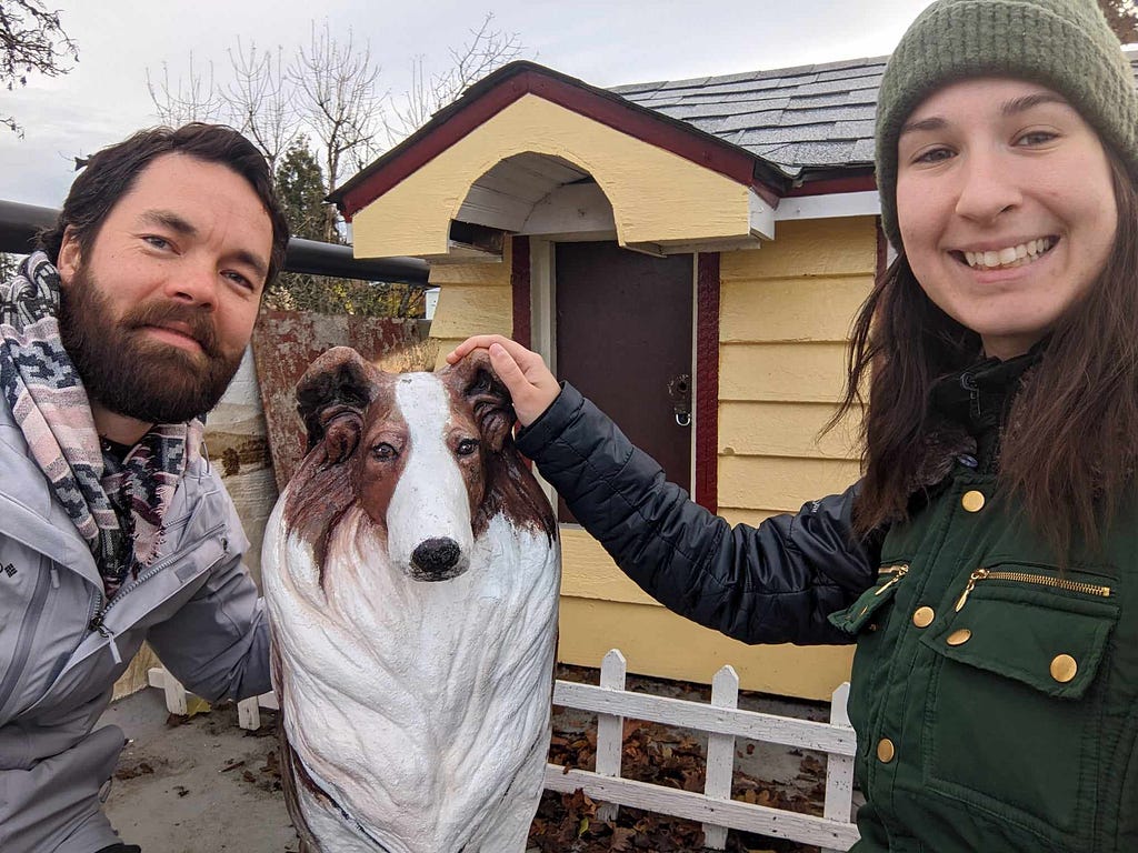 The author and her travel partner posing for a photo next to a dog statue, in front of a miniature dog house.