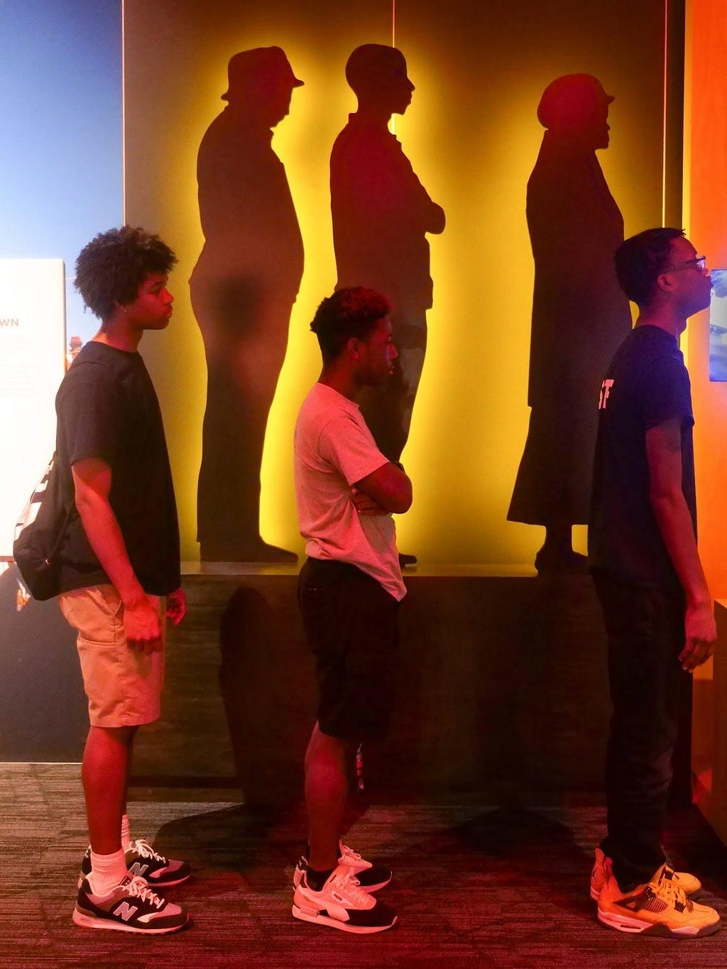Photograph of three Black teens standing sideways in front of a display that includes three black silhouettes against a yellow background.