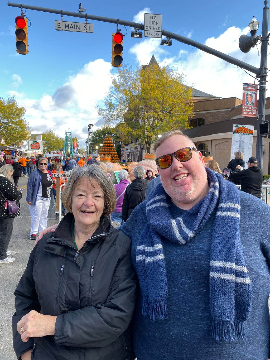 Two people looking at the camera smiling, standing on a busy street with a tower of pumpkins in the background