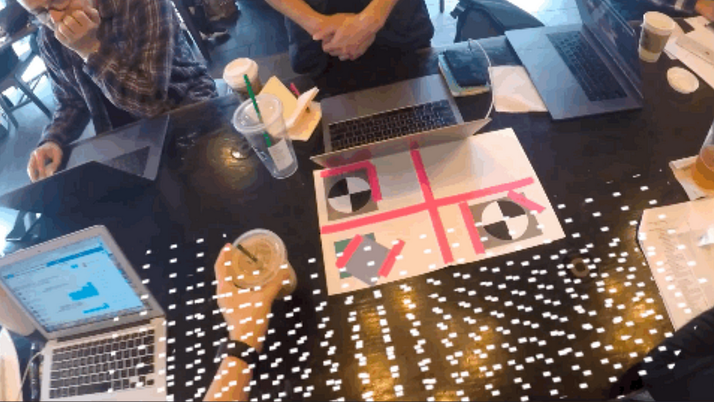 The image shows a group of people working around a table with laptops, drinks, and paper. A digital overlay of white dots or markers is visible across the table, suggesting the use of augmented reality or some form of 3D scanning technology in this collaborative workspace.