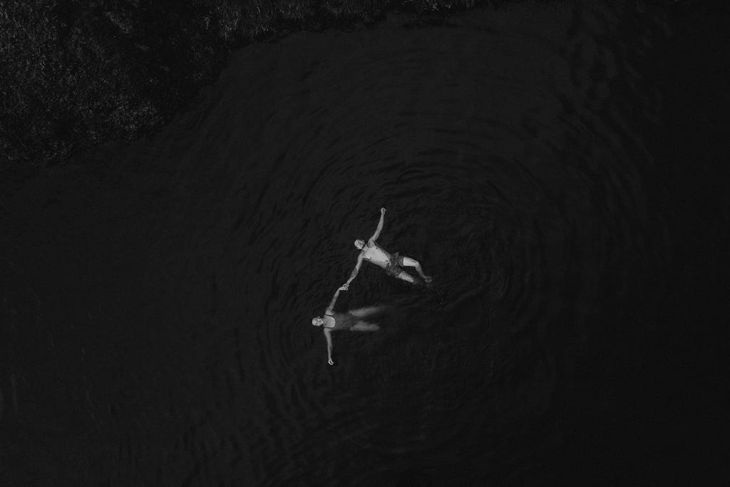 Man and woman holding hands, lying in the middle of dark waters.