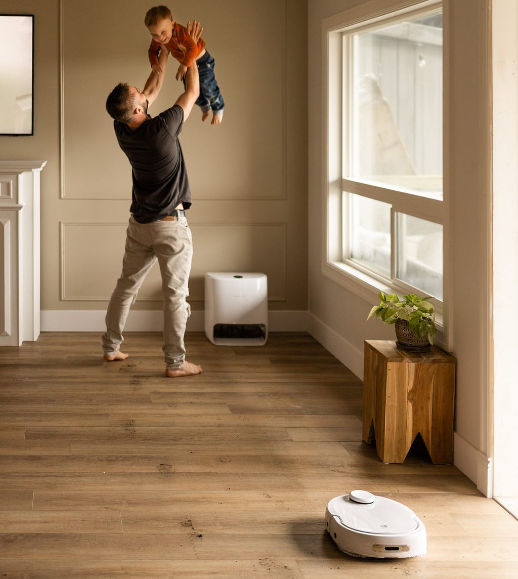 A man playing with his child as a cleaning robot cleans the floor