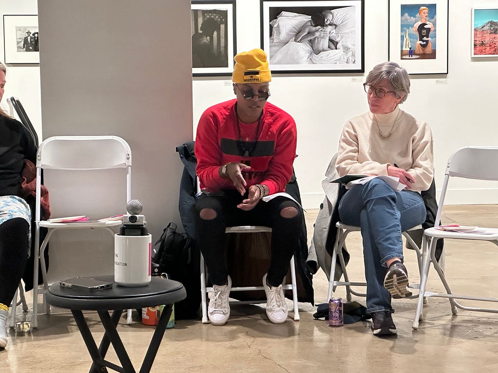 Two participants in a conversation, one a white appearing woman wearing jeans and a white sweater and the other a black appearing person wearing a red top, yellow cap, black pants and white tennis shoes.