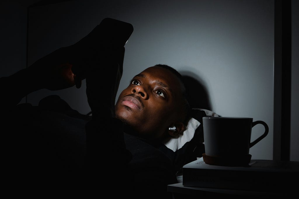 A person laying in the dark, their face illuminated by their phone with a cup of coffee on the night stand next to them