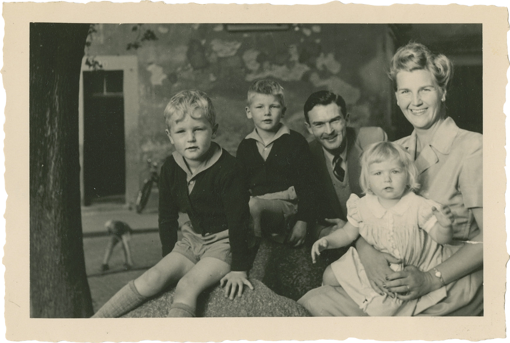 A smiling couple sits behind three children. Two boys are wearing sweaters, collared shirts, shorts and knee socks. The woman, with stylishly coiffed hair holds on her lap a very young girl wearing a dress with a white collar.