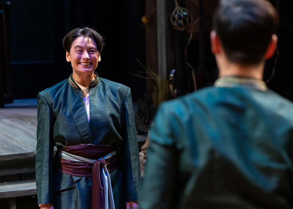 An actor with short dark hair and a teal coat smiles widely at another actor, who has their back to us but has similar short dark hair and a matching teal coat.