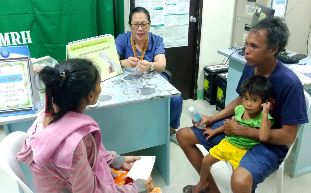 A midwife dressed in blue scrubs is seated at a desk and is speaking with a mother and father, who are accompanied by their child, about family planning options.