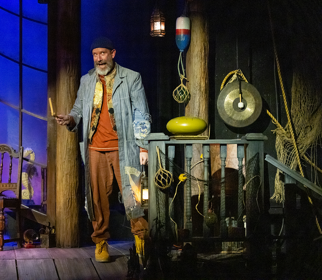 A performer in a denim coat with a beard sings near a gong and a tongue drum, while playing a ratchet noisemaker