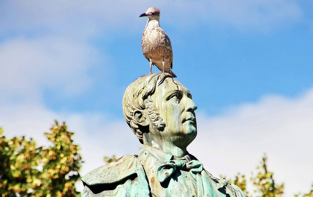 A seagull sits irreverently on top of a verdigris stained statue of a man, with a smattering of bird poo down its face