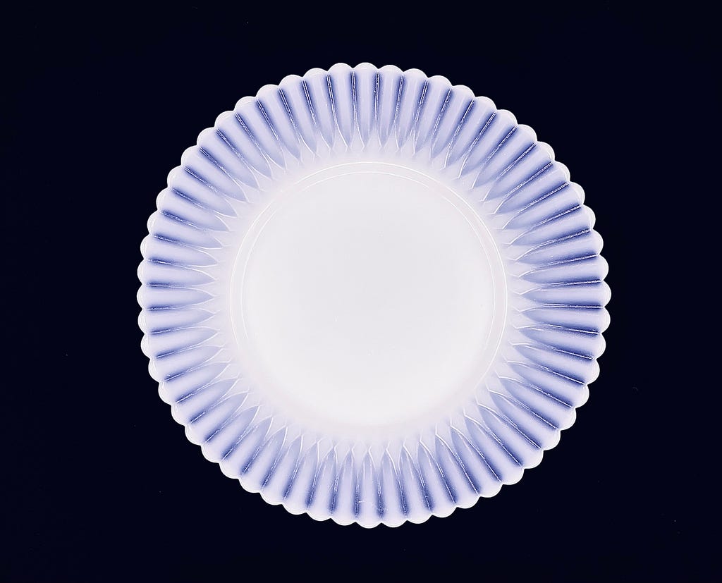 A top-down view of a Monax Petalware dessert plate against a black backdrop. The center of the plate is opaque while the decorative border at the outer edge is ribbed in a petal-like, sunburst pattern. Between the ribs is translucent purple against the black.