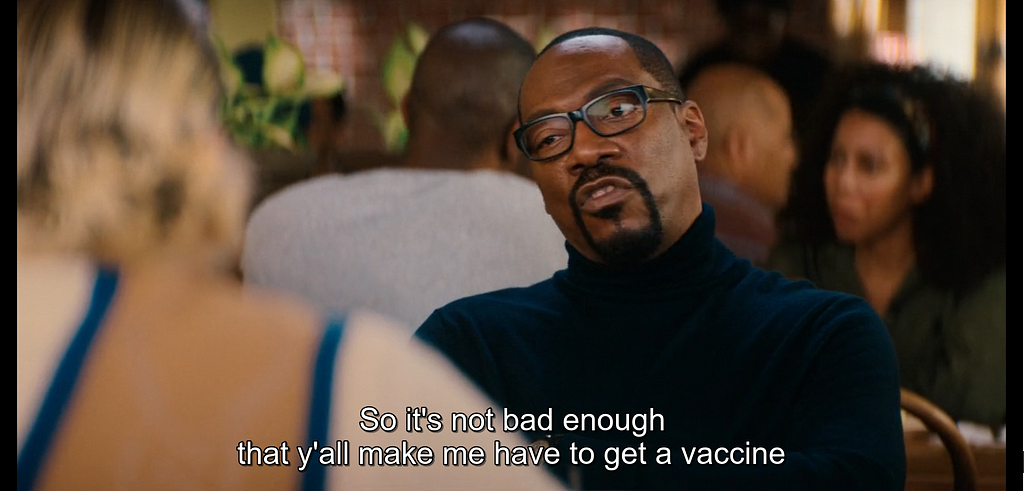 A man in a black turtleneck at a restaurant speaks sternly. Subtitles read, “So it’s not bad enough that y’all make me have to get a vaccine"