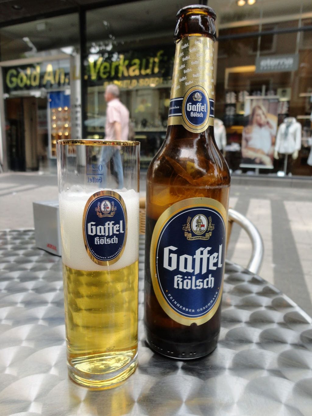 A glass of beer and a beer bottle with the label “Gaffel Kolsch” on a brushed metal outdoor table.
