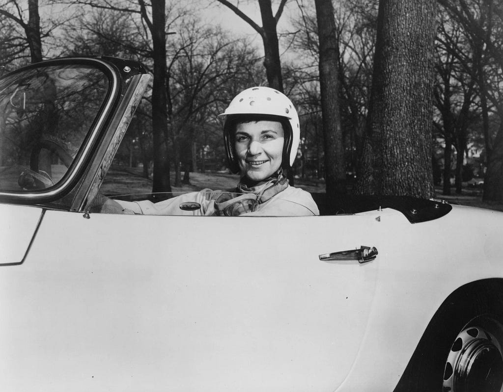 A woman wearing a spotted helmet smiles softly as she looks at the camera out of the window of a convertible.