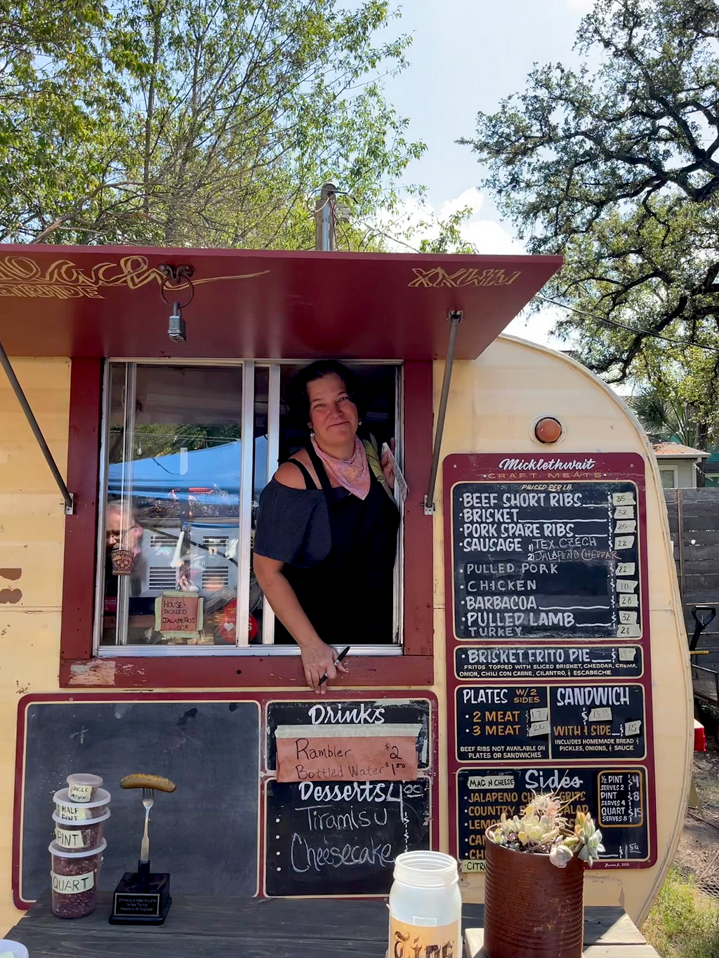 A woman smiling, leaning out of a caravan window with the menu on a blackboard next to her.