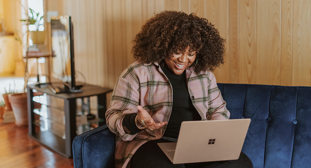 Photo by Surface on Unsplash — Woman with curly hair sitting on blue couch, smiling and gesturing to an open laptop computer on her lap