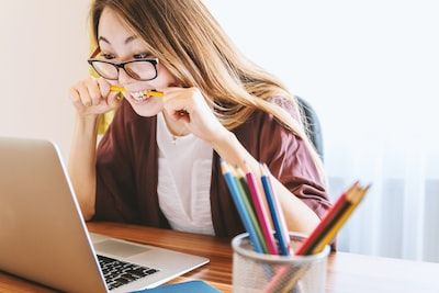 A girl wearing glasses an stressing about work is biting a pencil and staring at her computer at the same time