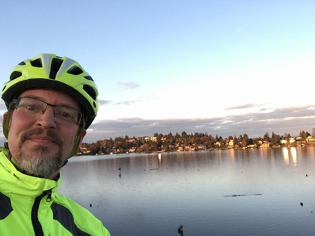 A man wearing a neon-yellow helmet and jacket stands in the foreground, with a lake and a wide city view off in the distant background.
