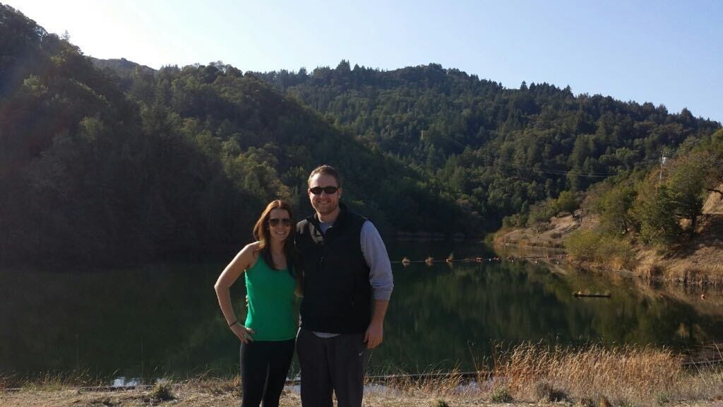 Alexa and her husband pose in front of a lake, with a green hillside in the background.