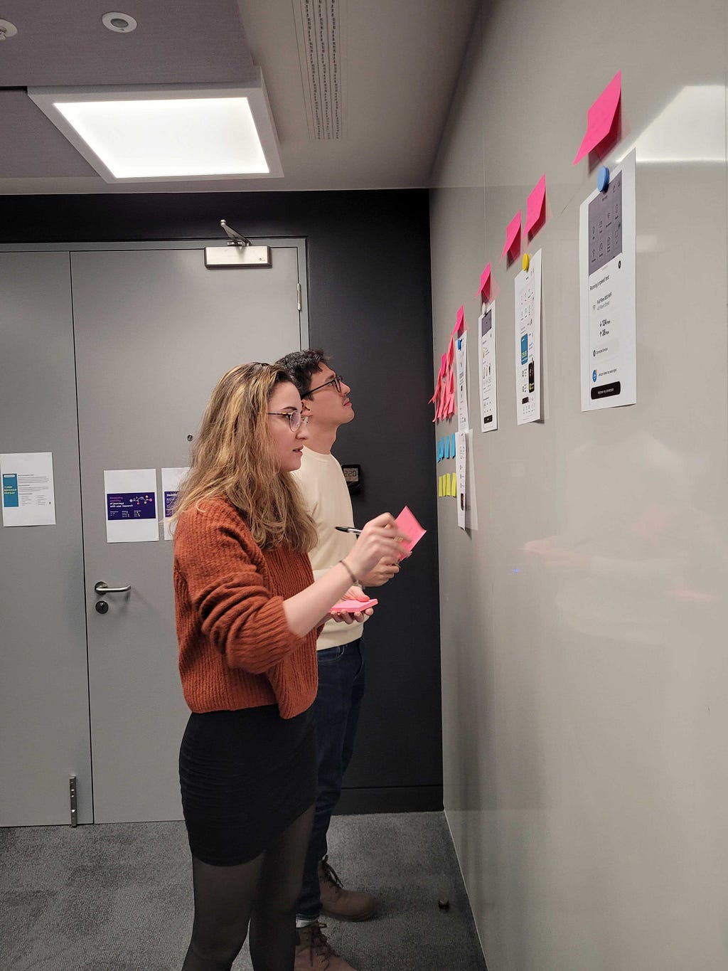 A male and female researcher stand facing a wall of post-its and print-outs of the digital design, making notes with more post-its against each print out of what they are learning.