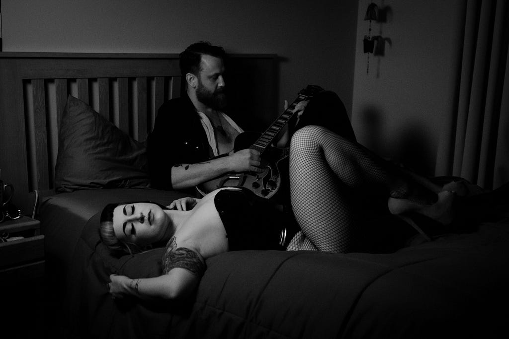 Black and white photo of a curvy, white woman in a black corset and fishnets, laying in bed next to a white, bearded man with a guitar