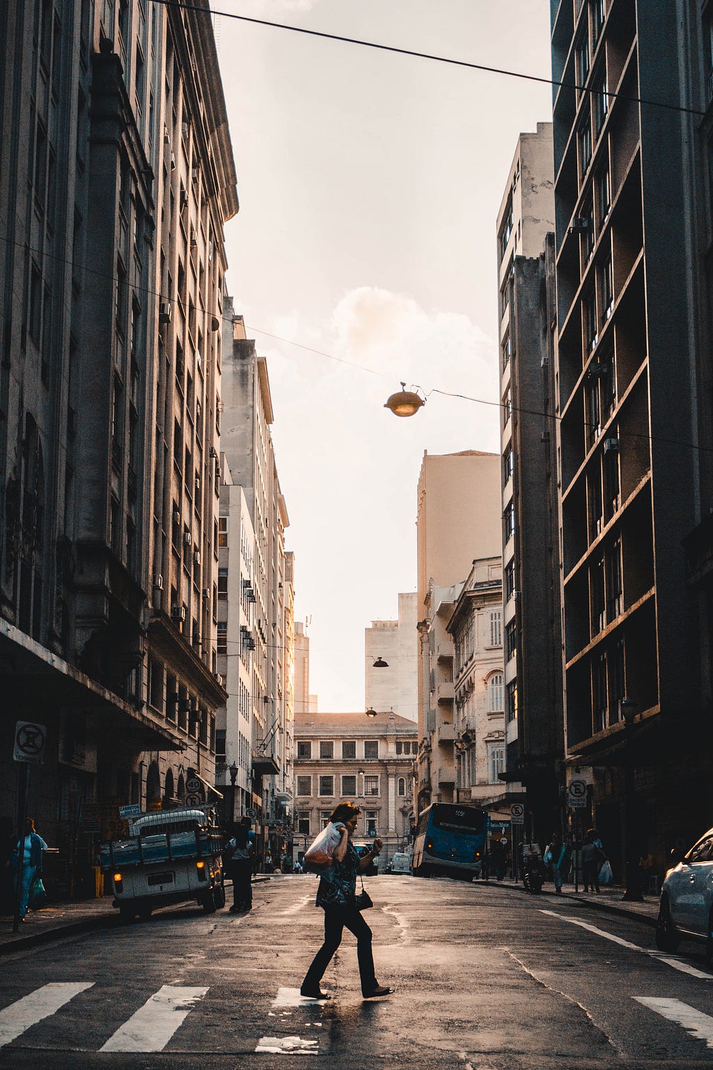 Rua do centro de São Paulo, cercada por inúmeros prédios antigos, com uma pessoa atravessando a rua, com a sacola nos ombros.