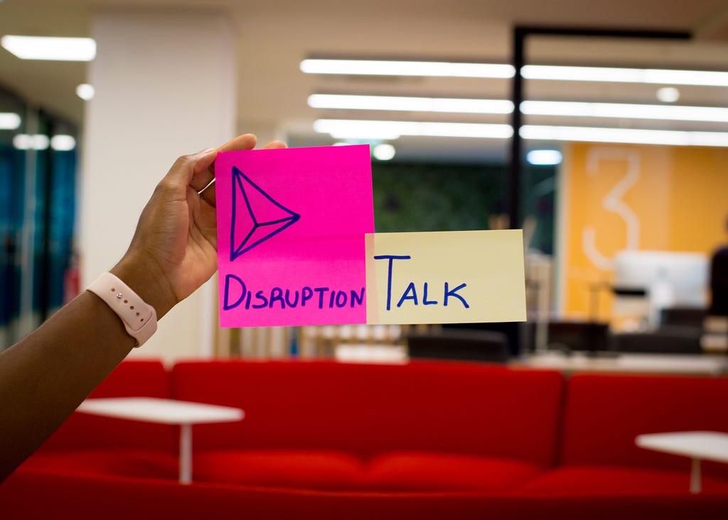 A hand holding up two Post-it notes. On one is written “Disruption” with a doodle of a pyramid. On the other is written “Talk.”