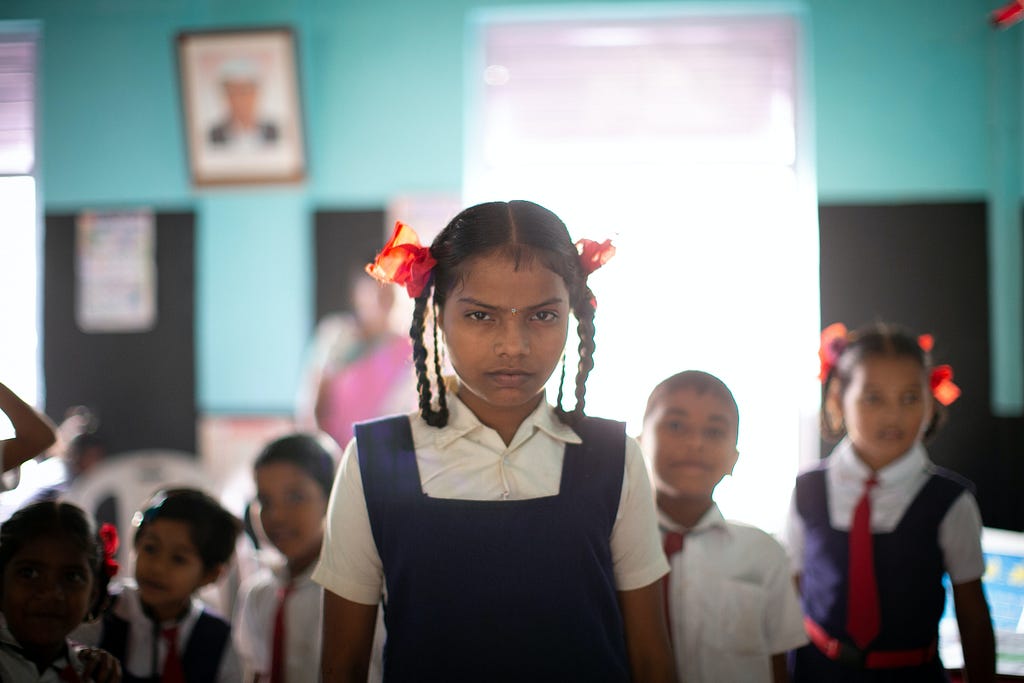 Picture of a girl in a school inform. Her hair is braided and she is looking intensely into the camera. In the background, there are five other children. Credit: Raj Rana via Unspalsh.