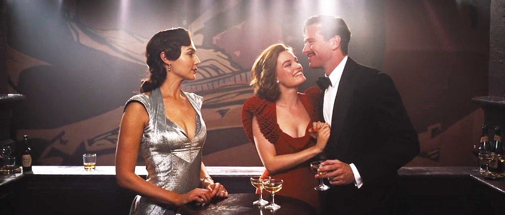 Gal Gadot in a silver dress (as Linnet Ridgeway) looks on as Emma MacKay in a red dress (as Jacqueline Bellefort) looks adoringly up at her fiance, Simon Doyle (Armie Hammer), who wears a tux. The clip is from Kenneth Branagh’s 2022 movie Death on the Nile.