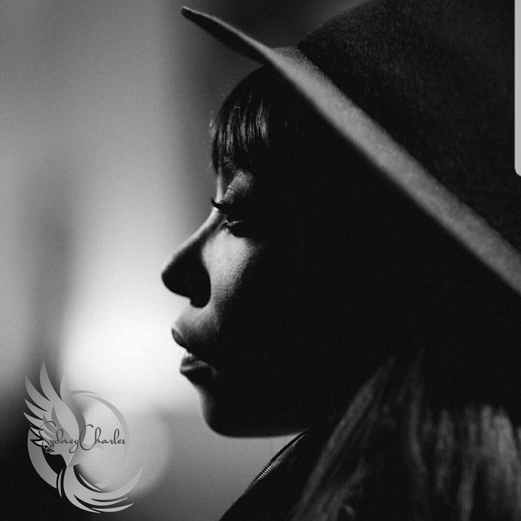This image features a black-and-white profile of a contemplative Black woman wearing a wide-brimmed hat. The lighting accentuates her nose and forehead, while shadows add depth to her thoughtful expression. A watermark with the name “Sydney Charles” suggests the author’s name in the bottom right corner.