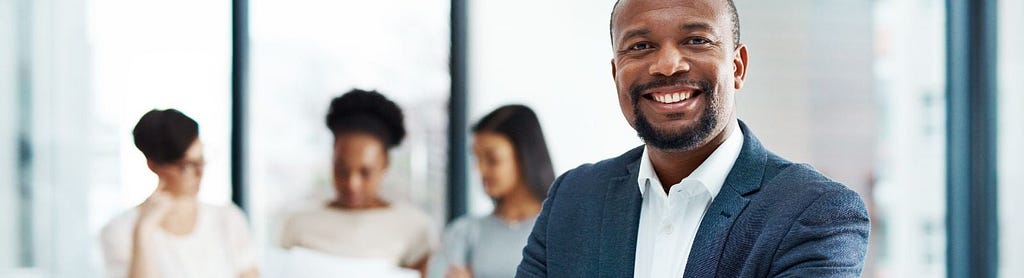Image of a man in the foreground smiling with 2 other coworkers in the background looking at a document