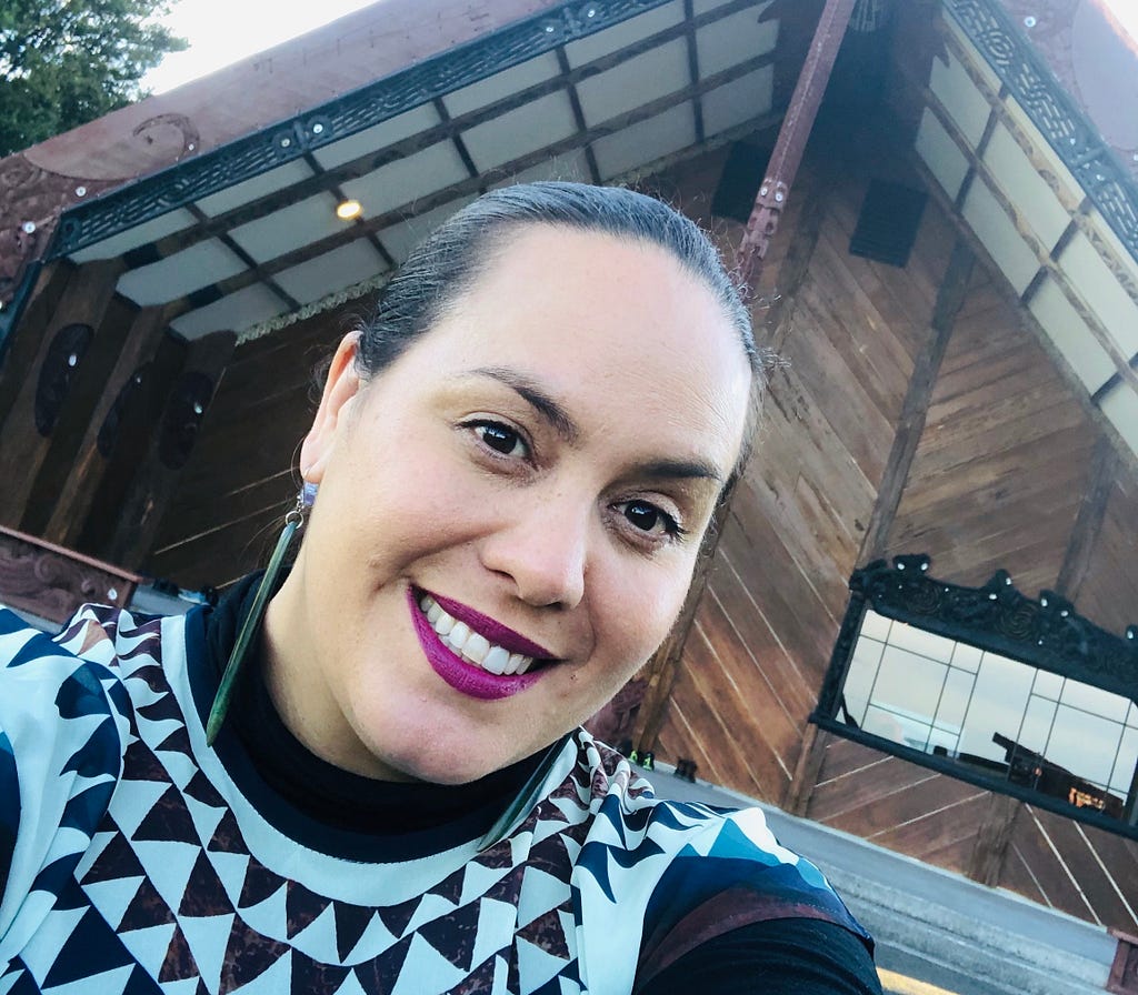 A selfie of the author, Ngapera Riley, a Māori woman. She is in front of a marae. She has pounamu earrings and a black and white top with a pattern inspired by nihoniho (triangle) designs.