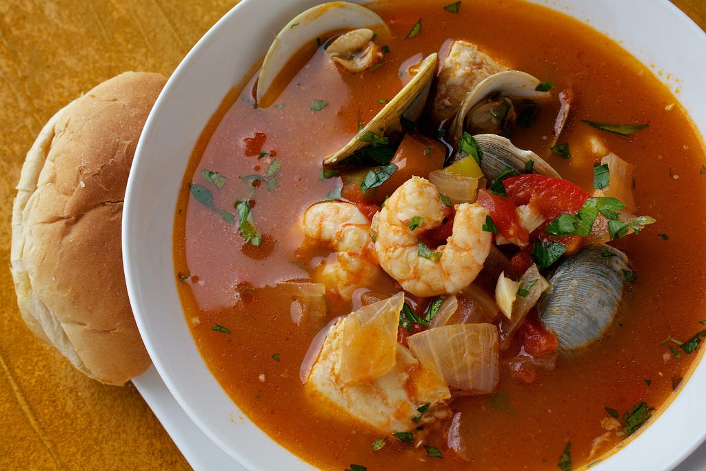 A bowl of soup with red broth, shrimp, crab, mussels, and veggies. Next to it is a roll of bread.