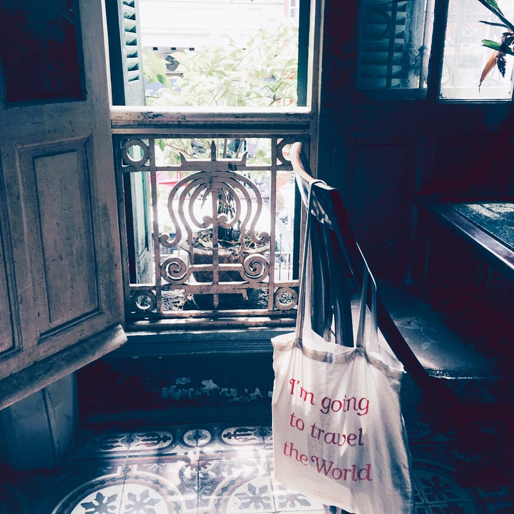 Image of a canvas bag that says “I’m going to travel the world”, on a chair looking out of a window. Cafe Thoughts Travelogue