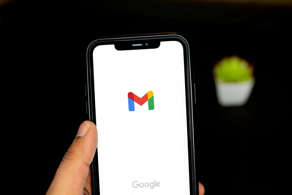 A hand holding a phone with a white background and the Gmail app screen open.