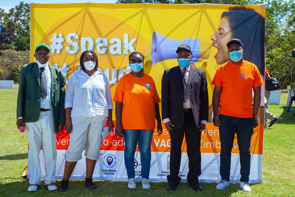 Group ohoto of two women and three men standing outside, wearing protective masks, and standing in front of a big and yellow photobooth where you can read “#Speak It Loud” with a woman holding a microphone