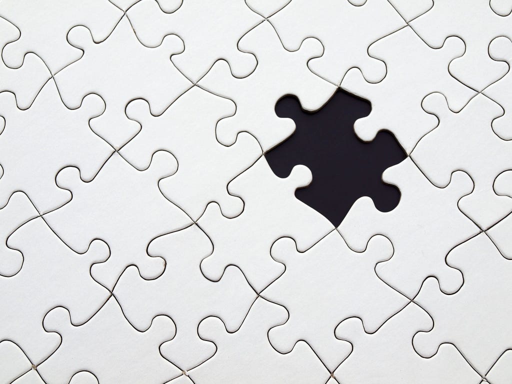 A photo of an all-white jigsaw puzzle with one piece missing, revealing a black background.