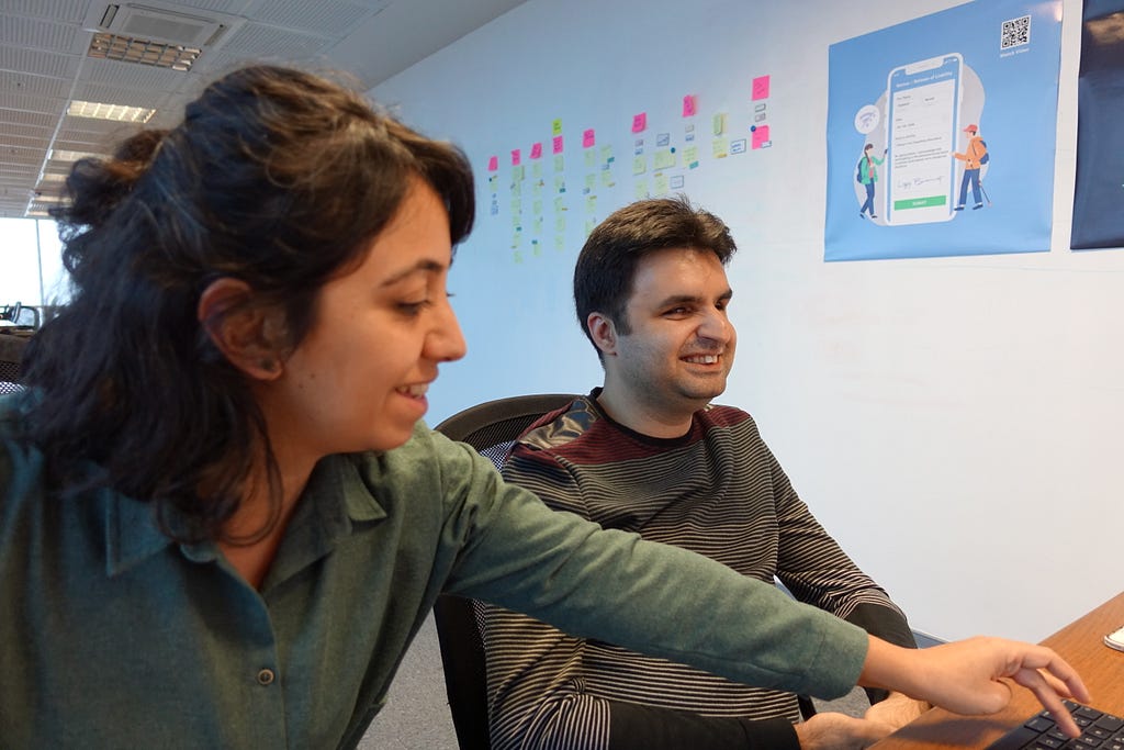 A photo of user researcher Gokce and Cagrı having a usability test next to a white wall with post-its and posters on it.