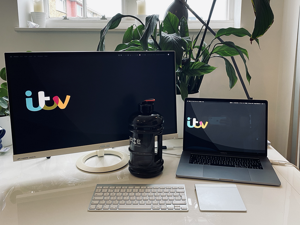 A laptop and a monitor sit on a white table. The ITV logo appears on each screen, and a peace lily sits behind them.