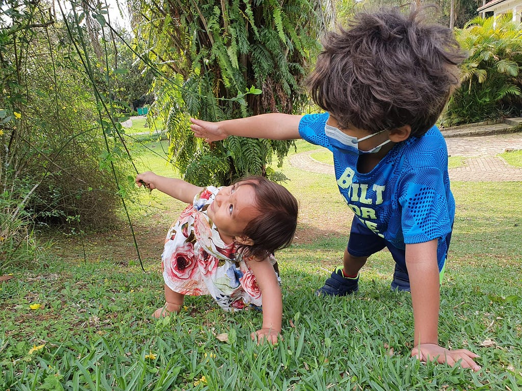 Foto de uma bebê e um menino escalando um morrinho verde. Ambos estão com uma mão no chão e outra erguida. A bebê sorri.