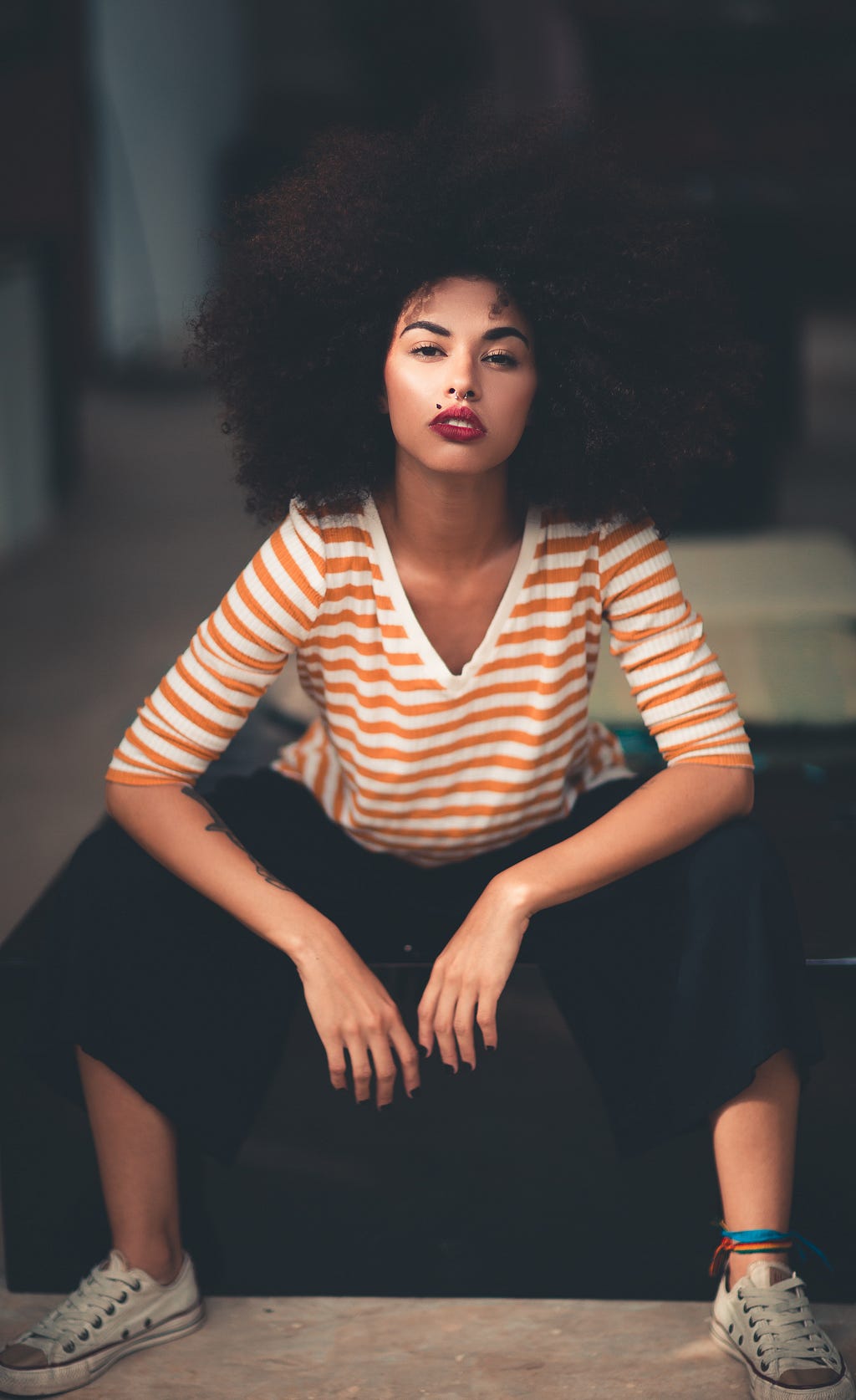 A black woman with an afro is sitting down. She is wearing a striped white and orange shirt with black pants. Her elbows rest on her knees.