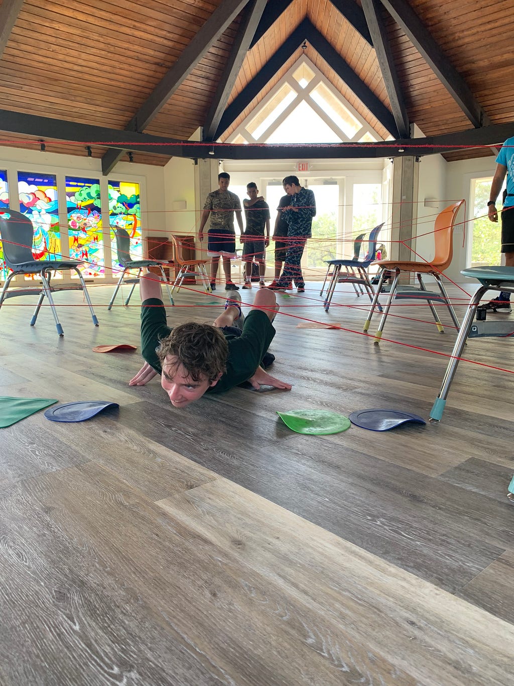 camper slides across the floor trying to avoid touching a yarn web above them. The yarn web is held up by various chairs in the sanctuary. Three other campers are in the background waiting for their turn