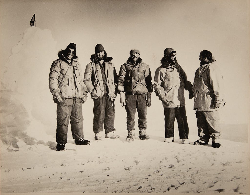 Sepia toned image of 5 men standing by a snow pile in Antarctica. All are wearing thick coats and trousers, mittens and hats to protect from the cold. 3 men are smiling at the photographer, 2 are facing each other.