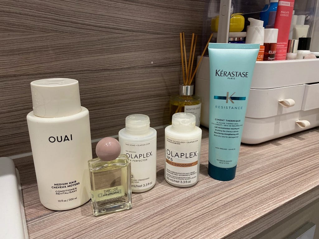 Image of 5 haircare products. The first is a conditioner by OUAI in a white bottle. The second is a small glass bottle with a purple circular screw lid. The bottle says ‘Hair Oil’. The 3rd and 4th are Olaplex bottles and the last is a Kerastaste Ciment Thermique bottle. It is blue and taller than the other bottles.