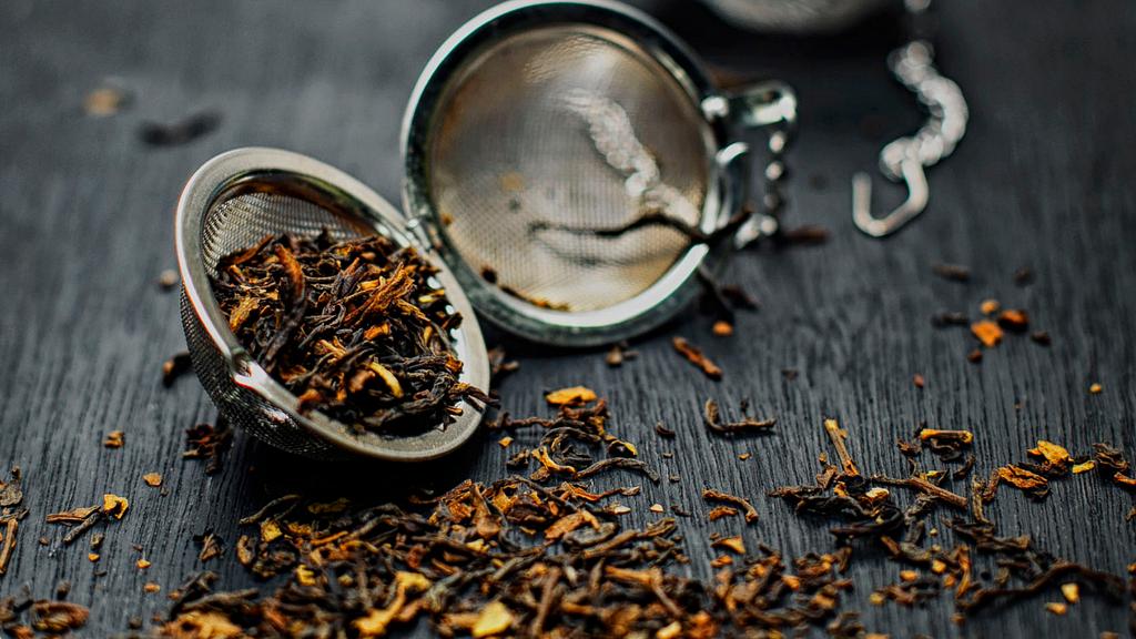 An open metallic mesh tea ball on top of a dark wooden table. An assortment of black tea leaves sit in half of the ball. The rest of the leaves are scattered around the table in front of it.