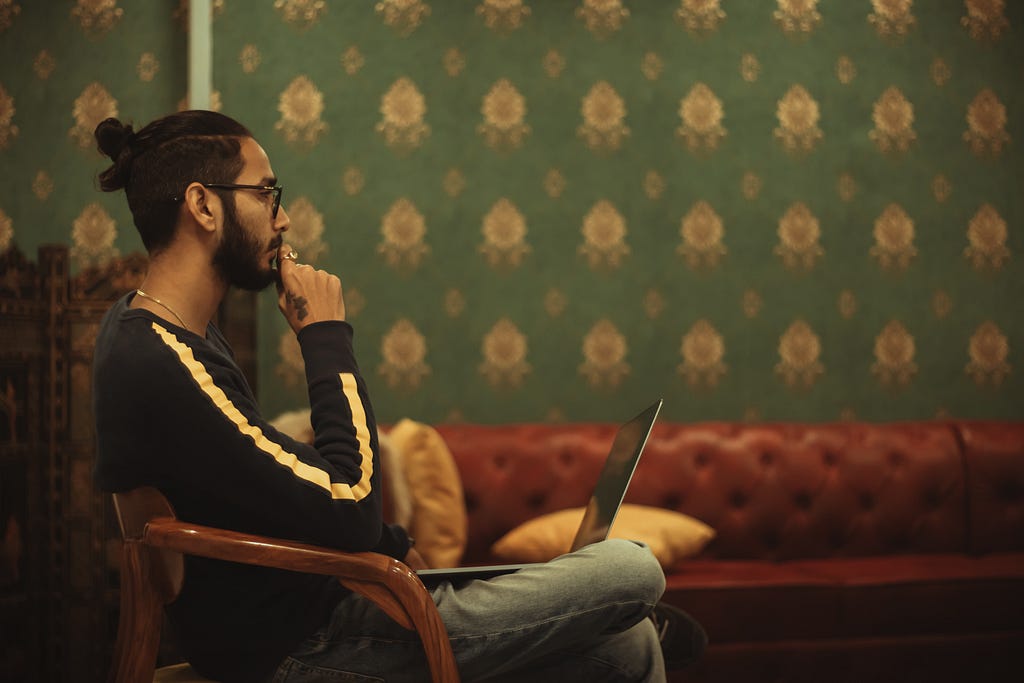 a person sits in a wooden chair. they are wearing glasses, they have a laptop in their lap and they look thoughtful.