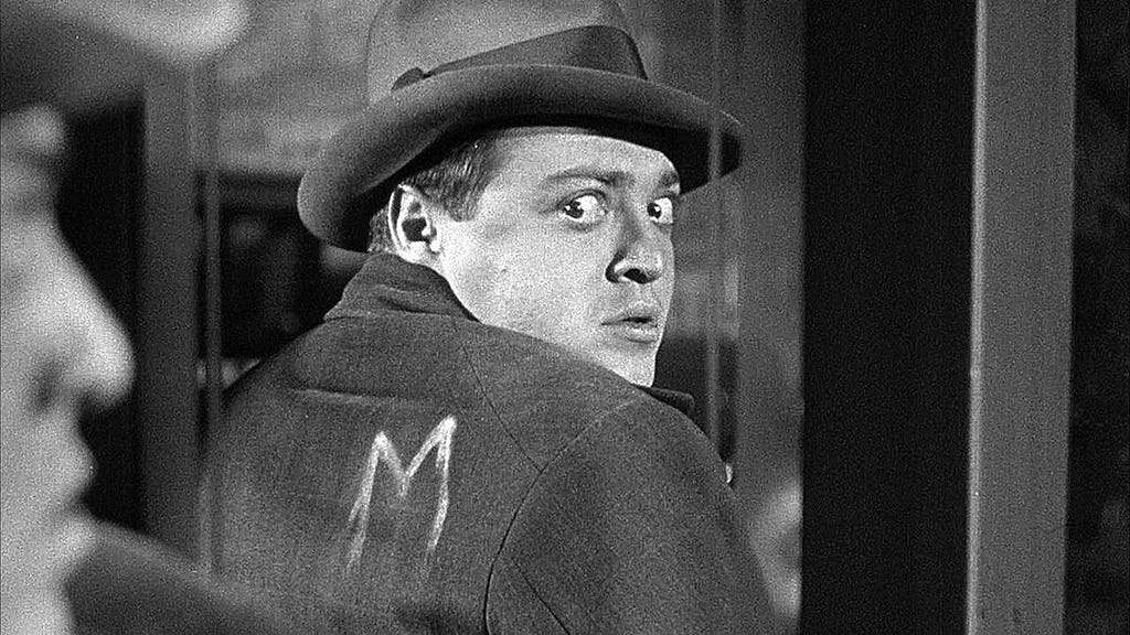 A man in a hat looks back at himself in a mirror with the letter M scrawled on his back.