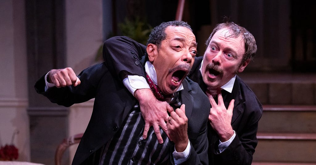 A man with curly dark hair wearing a black jacket and striped vest looks wide-eyed while another man with a moustache and soul patch puts his right arm around him in a gesture of friendship.